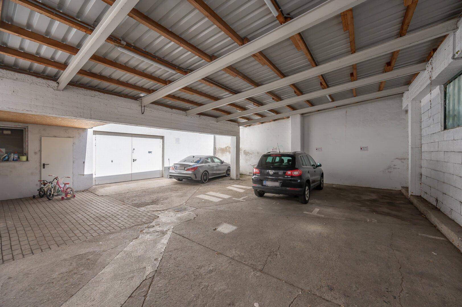 Überdachter Parkplatzbereich mit freiliegenden Holzbalken und zwei abgestellten Fahrzeugen.