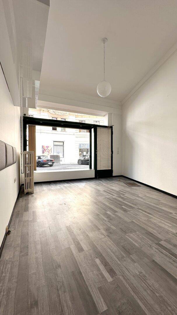Geräumiger Geschäftsraum mit hohen Decken, großem Fenster und hellem Laminatboden, bietet viel Licht.