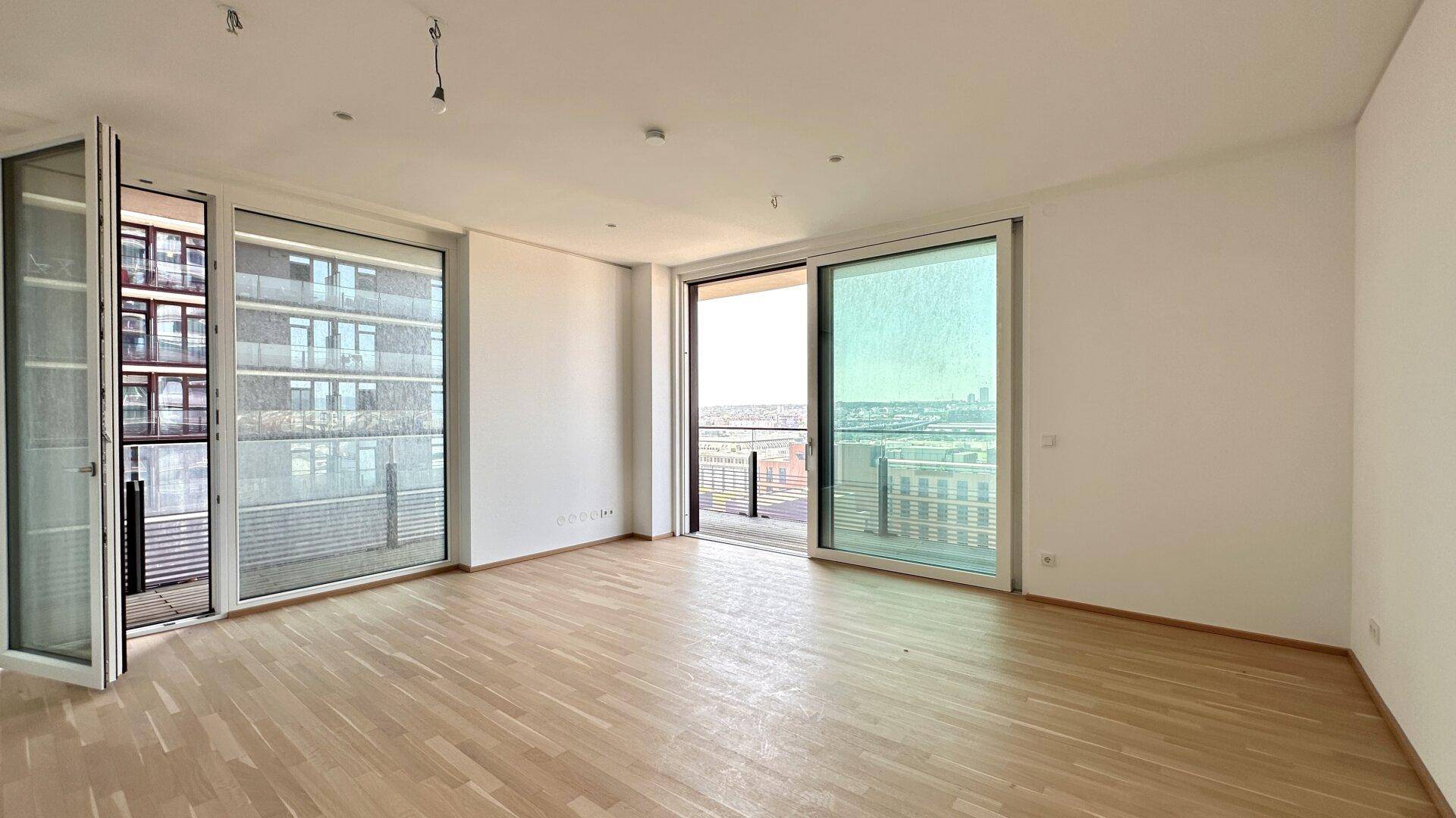 Helles, leeres Wohnzimmer mit großen Fenstern, Holzboden und Zugang zum Balkon.