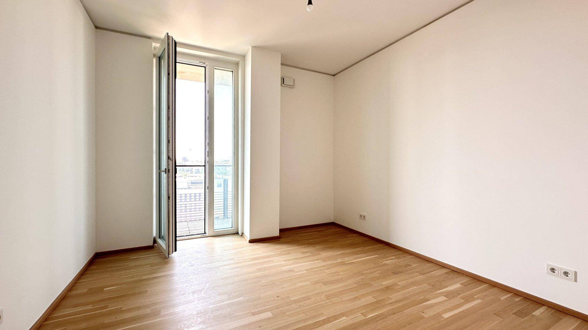 Leeres Schlafzimmer mit Holzboden und bodentiefem Fenster, das viel Tageslicht hereinlässt.