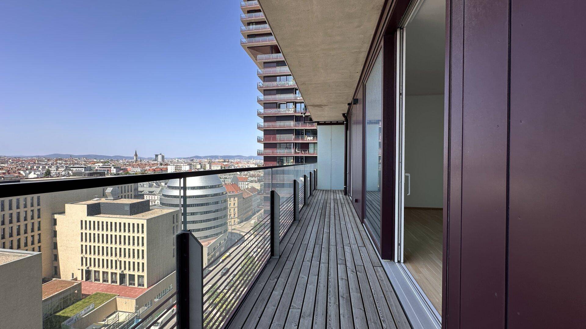 Langer Balkon mit Holzboden, Glasgeländer und weitem Blick über die Stadt.