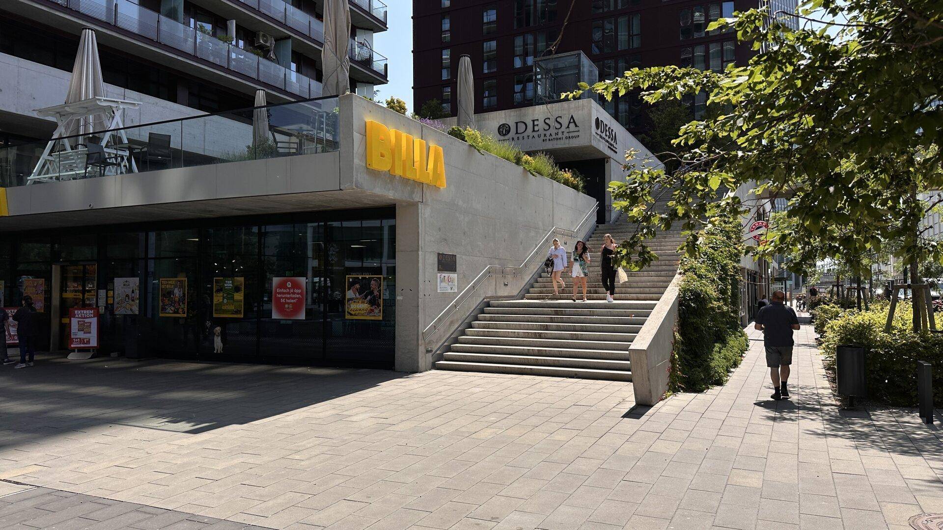 Eingangsbereich eines modernen Gebäudes mit Restaurant und Außentreppe in urbaner Umgebung.