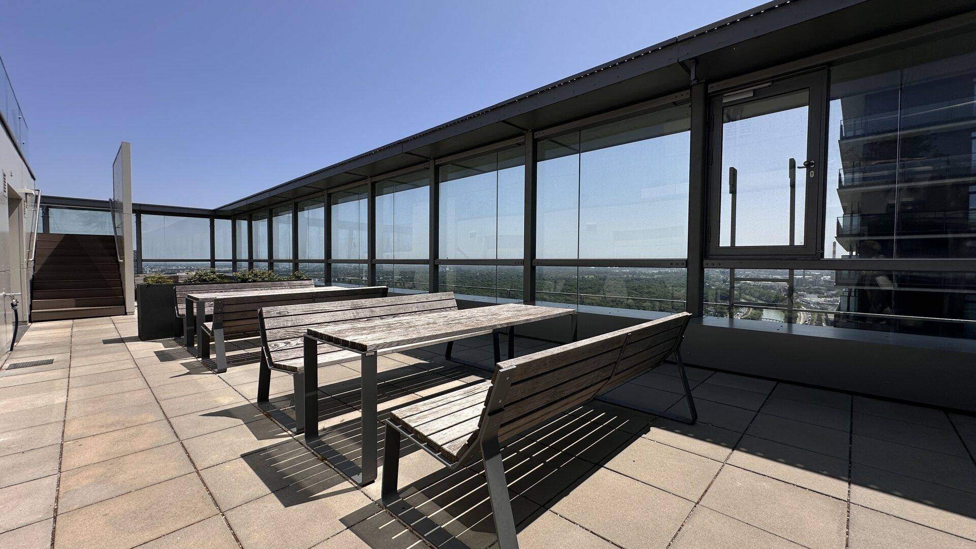 Gemeinschaftliche Dachterrasse mit Holzbänken und Tischen, umgeben von Glaswänden und Stadtblick.