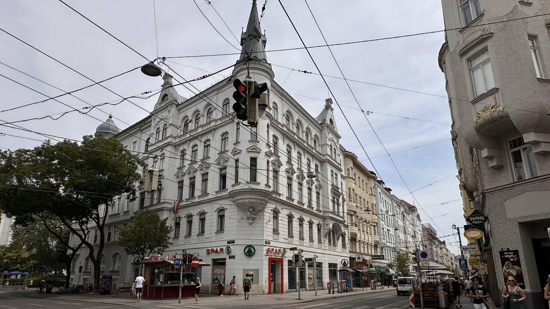 Historisches Eckgebäude mit vielen Fenstern und Geschäften im Erdgeschoss an einer belebten Straße.