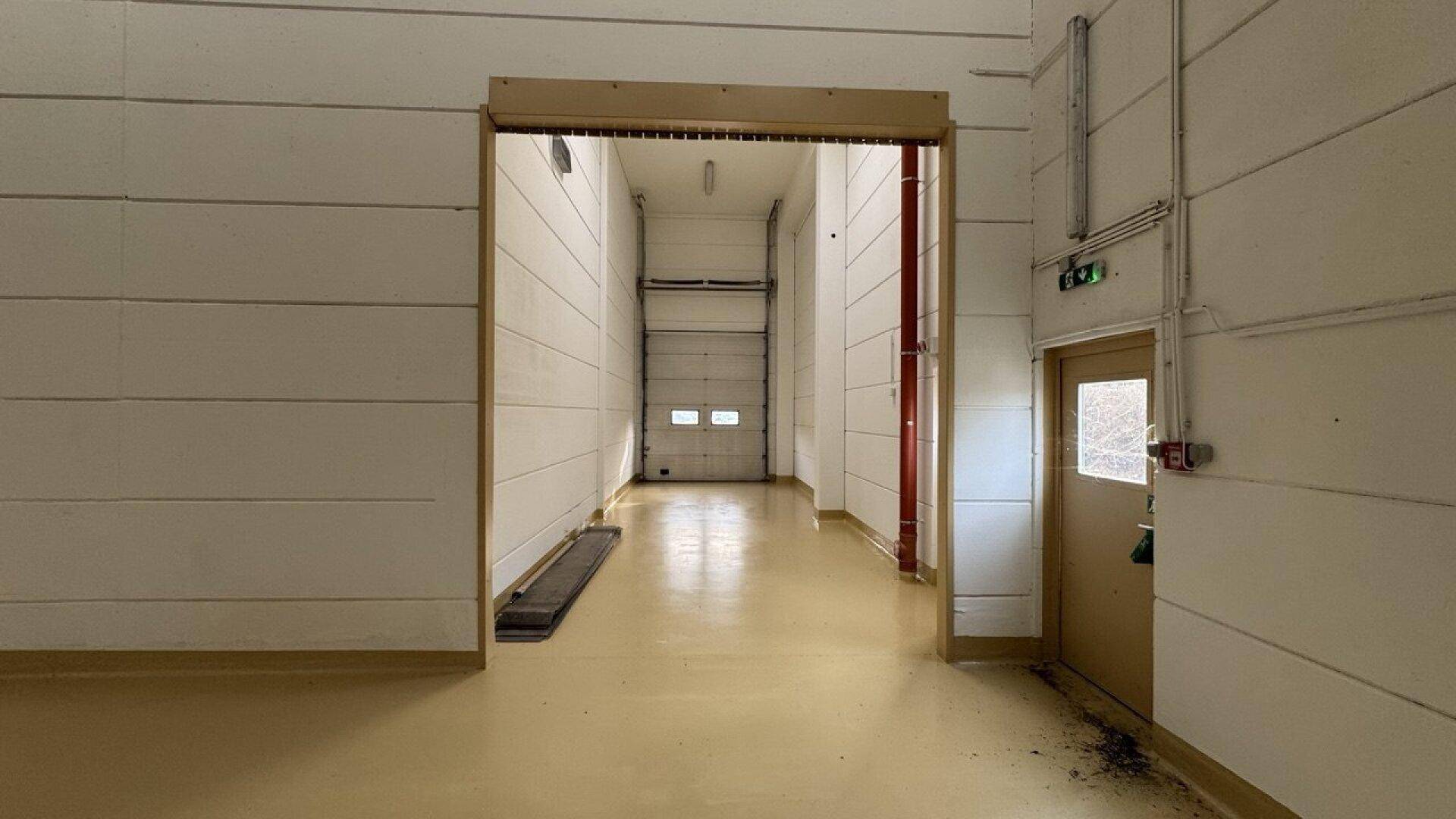 Langer Flur in einer Industriehalle, der zu einem großen Rolltor und einer Seitentür führt.