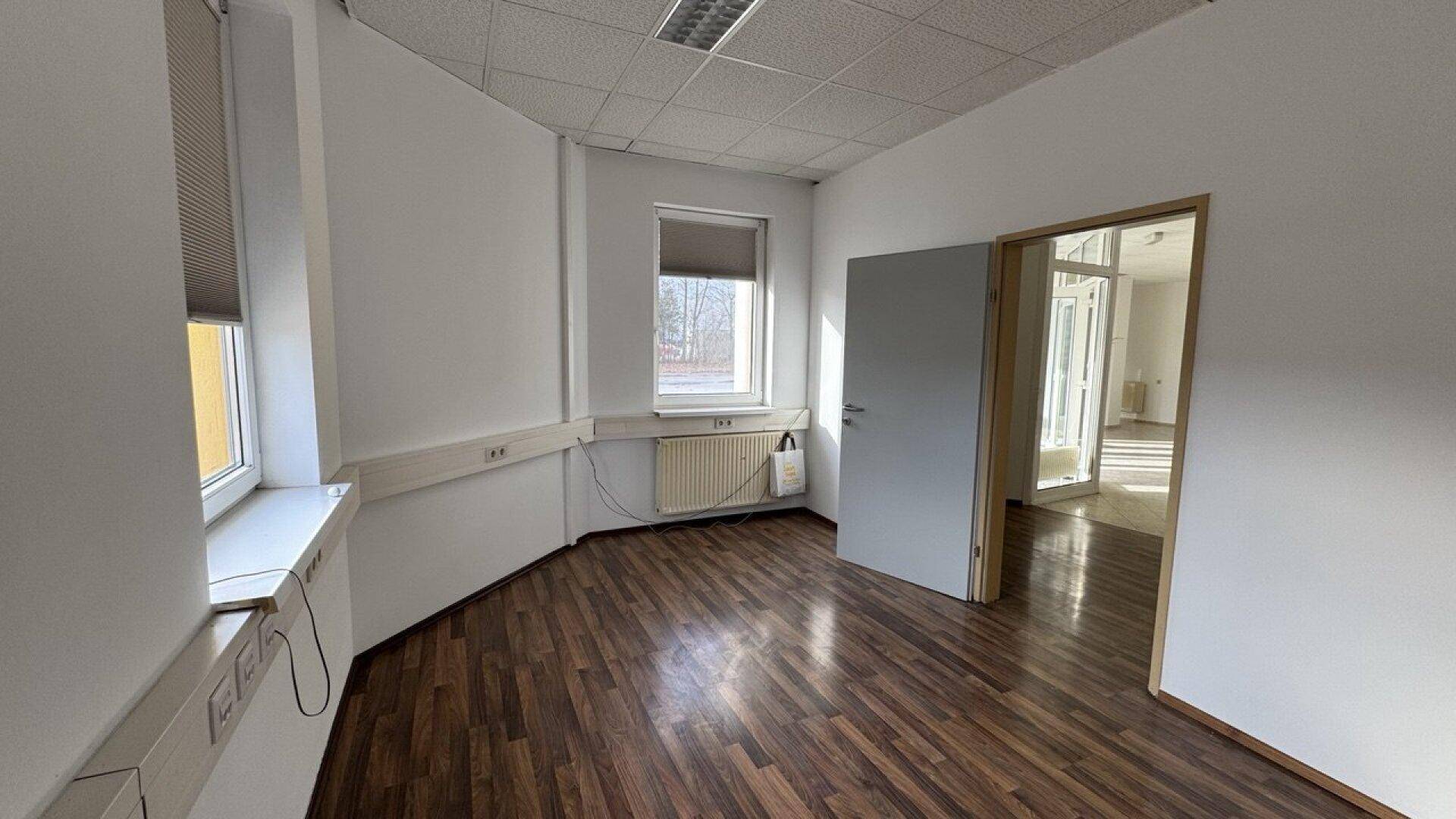 Helles Büro mit dunklem Holzboden, einem Fenster mit Jalousie und einer Tür zu einem weiteren Raum.