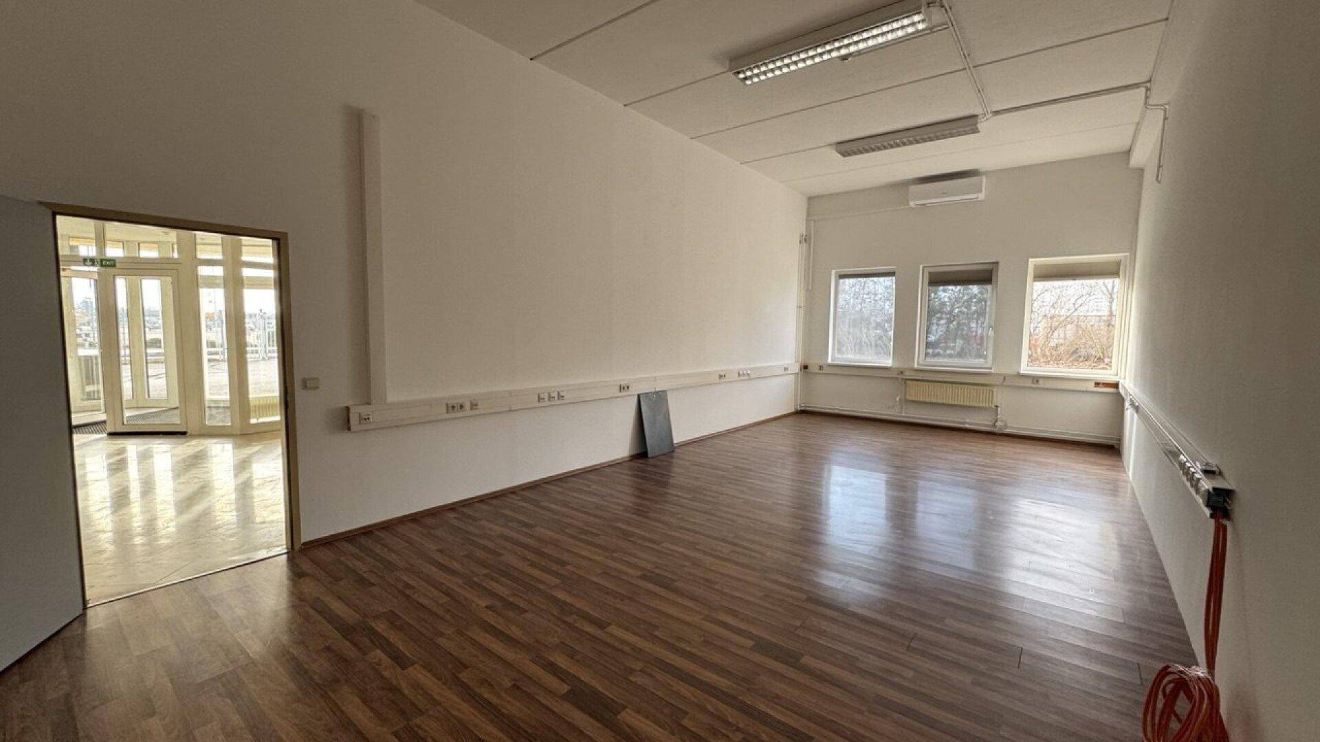 Geräumiges Büro mit dunklem Holzboden, weißen Wänden und Blick in einen hellen Nebenraum.