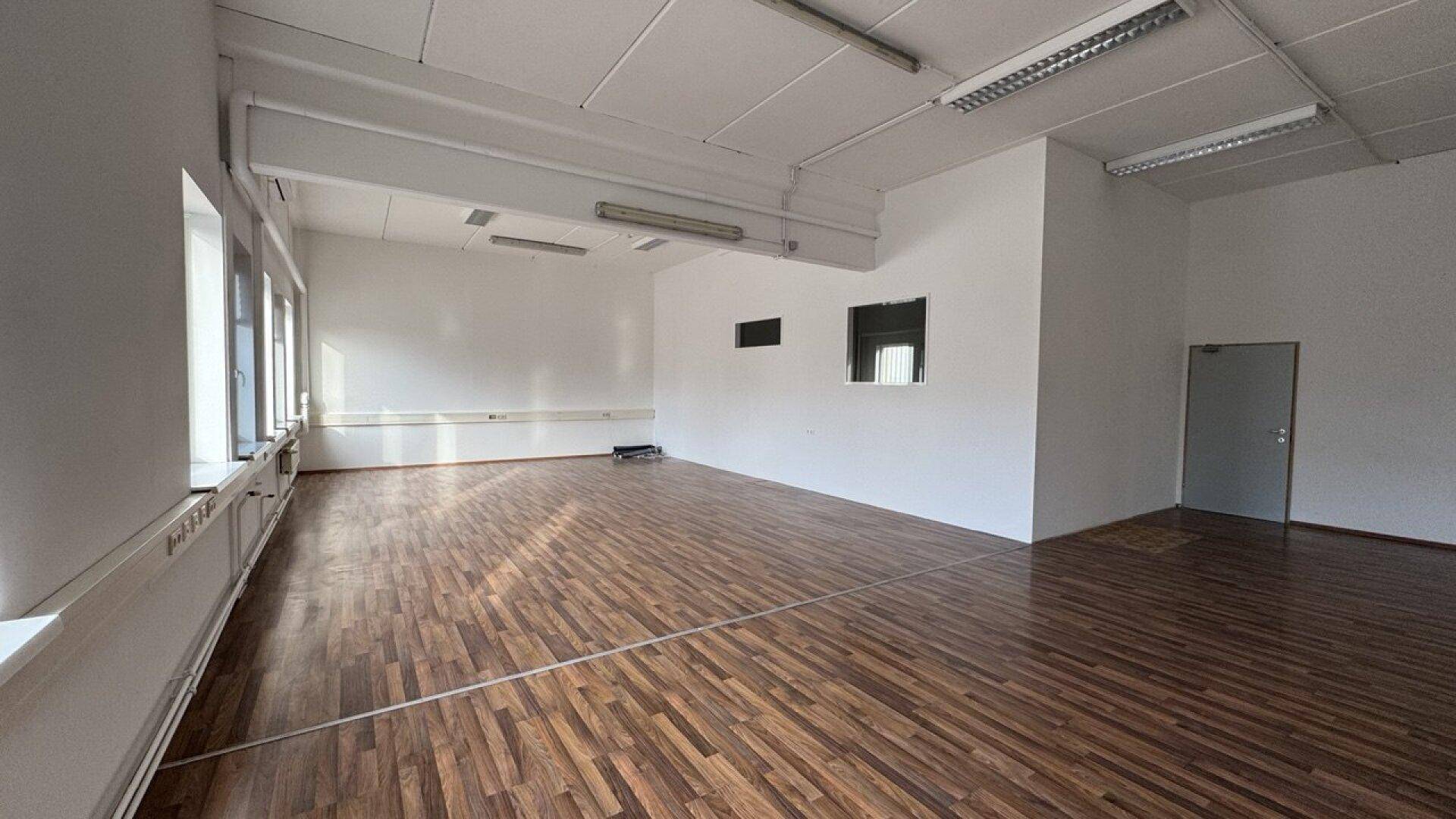 Großes Büro mit dunklem Holzboden, vielen Fenstern und einer schlichten, funktionalen Einrichtung.