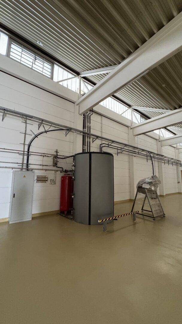 Technikbereich in einer Lagerhalle mit sichtbaren Rohren, Schaltschränken und einem großen Wassertank.