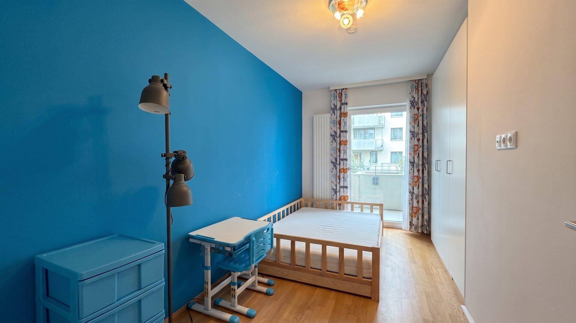 Kinderzimmer mit blauer Wand, Holzbett und Schreibtisch, sowie Zugang zum Balkon.