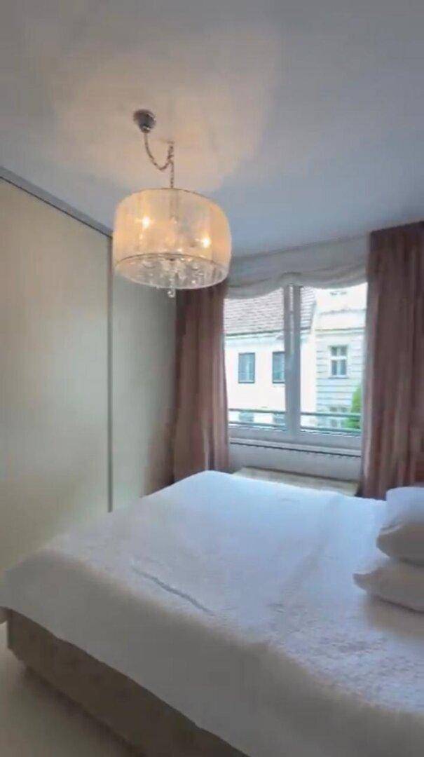 Helles Schlafzimmer mit großem Bett, Fenster und einer eleganten Deckenleuchte.