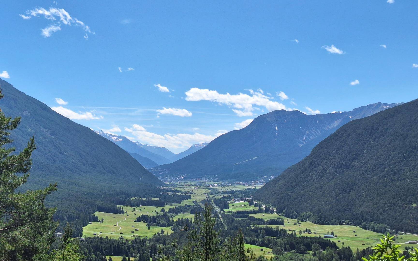 Weitläufiger Panoramablick über ein grünes Tal mit Wäldern und einem Dorf, umgeben von majestätischen Bergen.