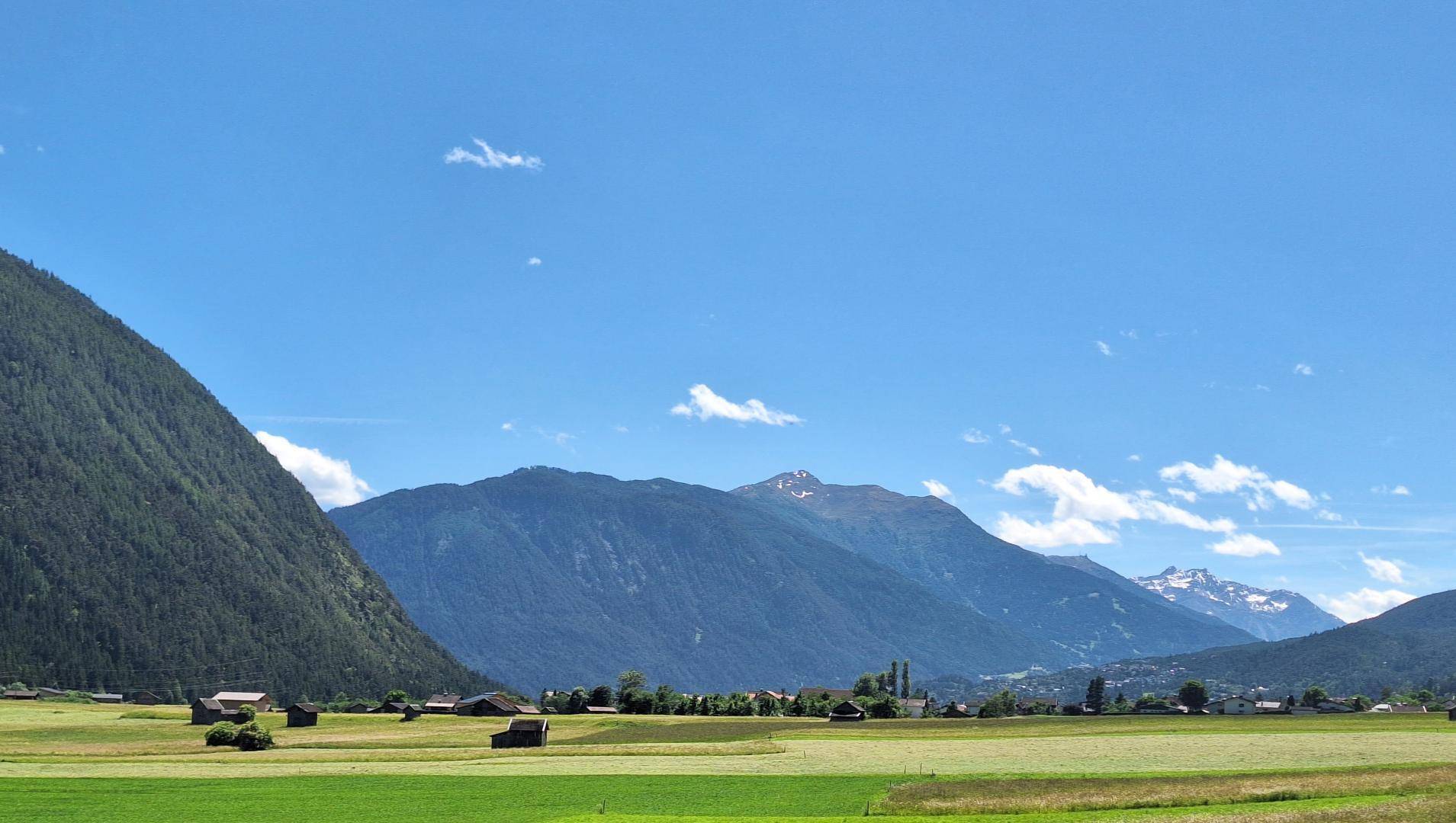 Idyllische Landschaft mit weiten grünen Feldern, kleinen Häusern und einer beeindruckenden Bergkulisse im Hintergrund.