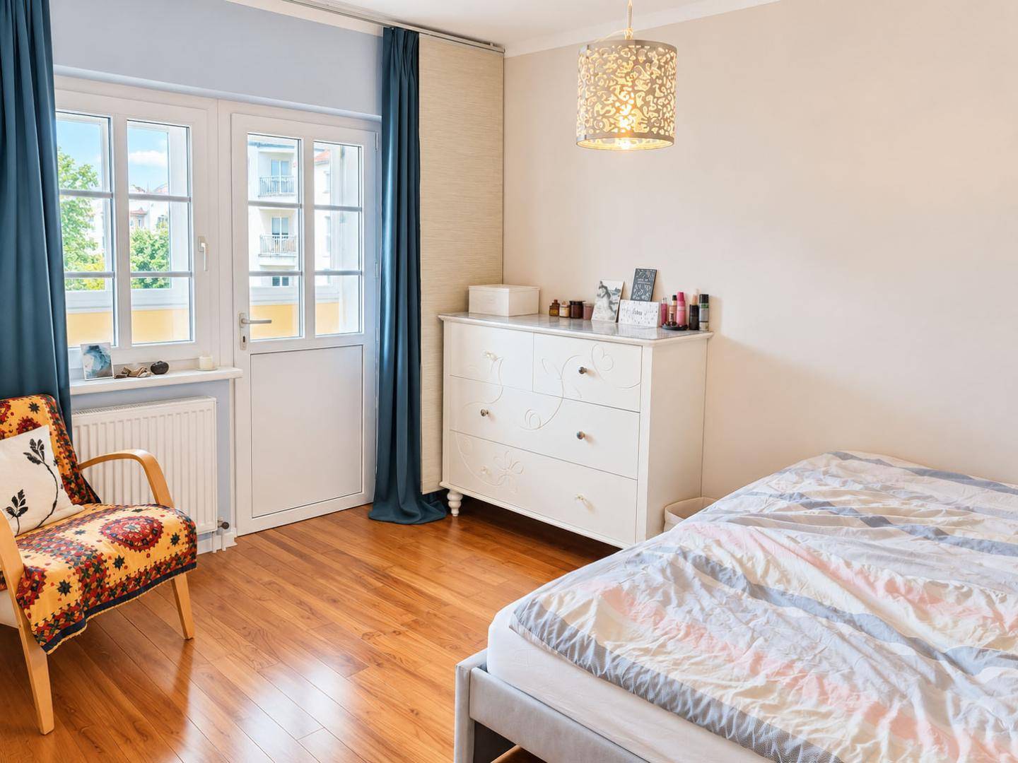 Helles Schlafzimmer mit Holzboden, Kommode und Zugang zu einem Balkon.