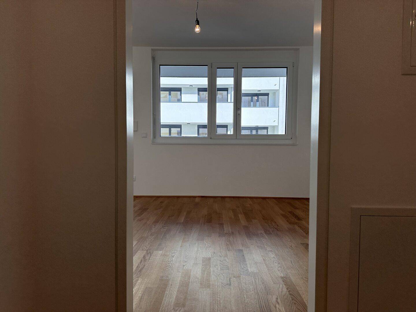 Blick vom Flur in ein helles Zimmer mit Parkettboden und großem Fenster.