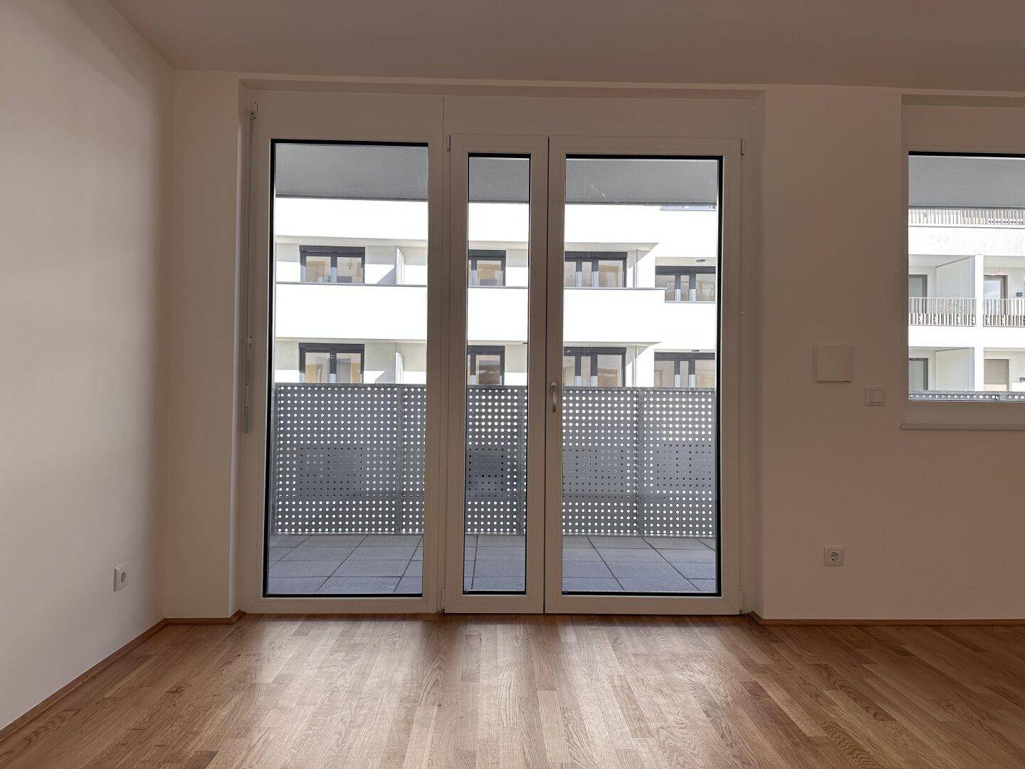 Geräumiges Zimmer mit Parkettboden und großen Fenstern zum Balkon.