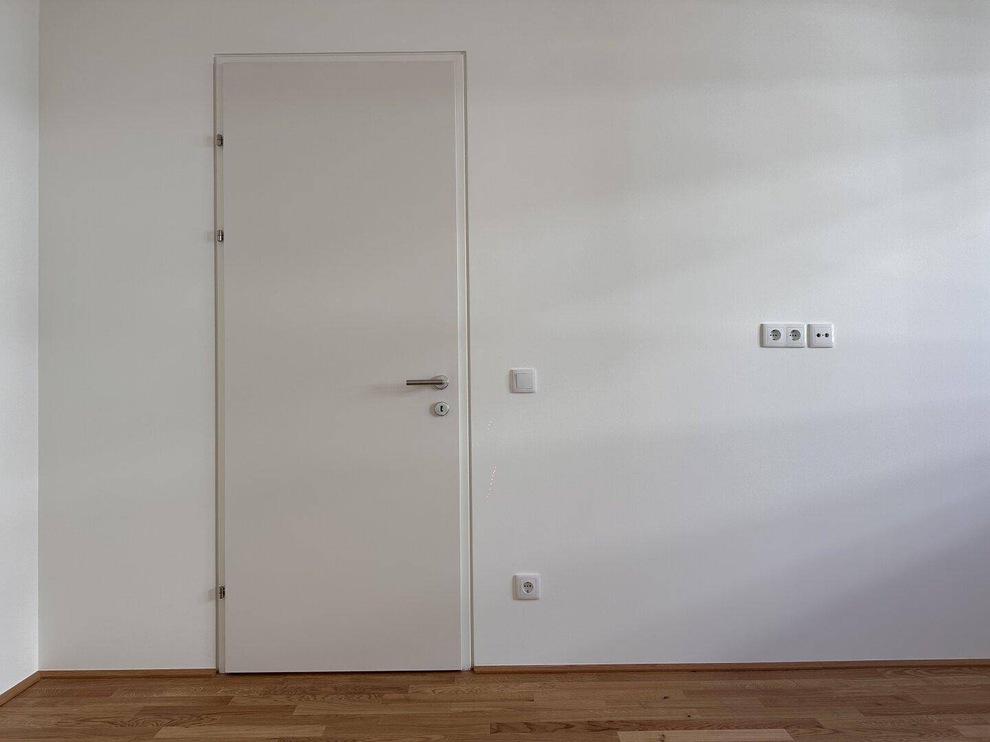 Helles Schlafzimmer mit Parkettboden und einer schlichten weißen Tür.