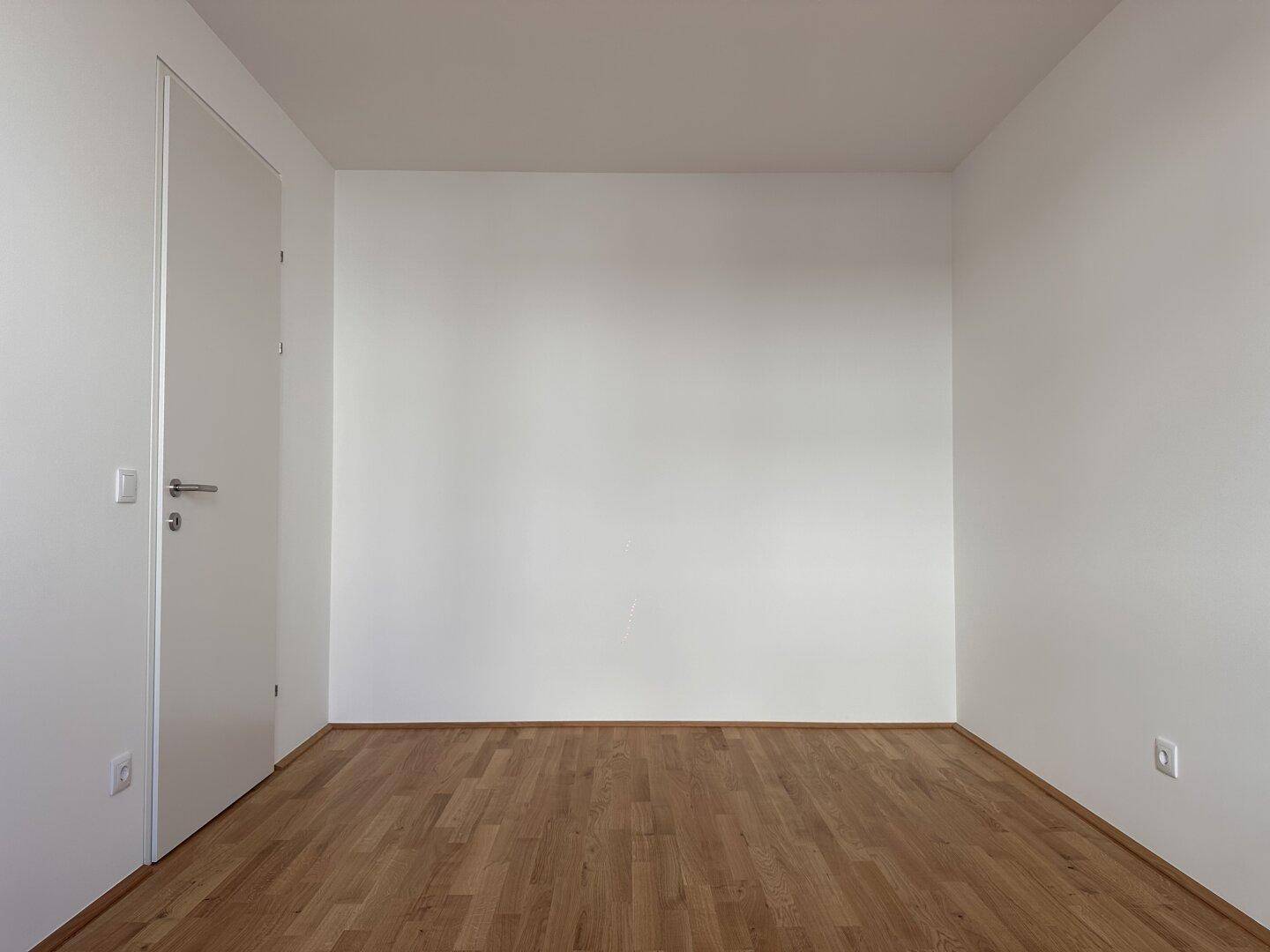 Helles und leeres Zimmer mit Parkettboden und weißen Wänden, bereit zur individuellen Gestaltung.