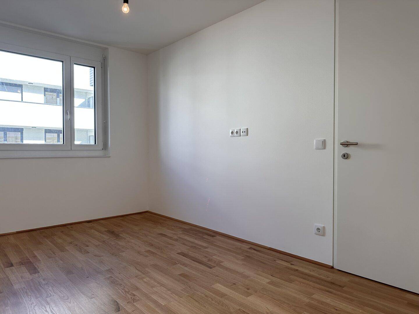 Helles Zimmer mit Parkettboden, Fenster und moderner Tür, ideal als Schlaf- oder Arbeitszimmer.