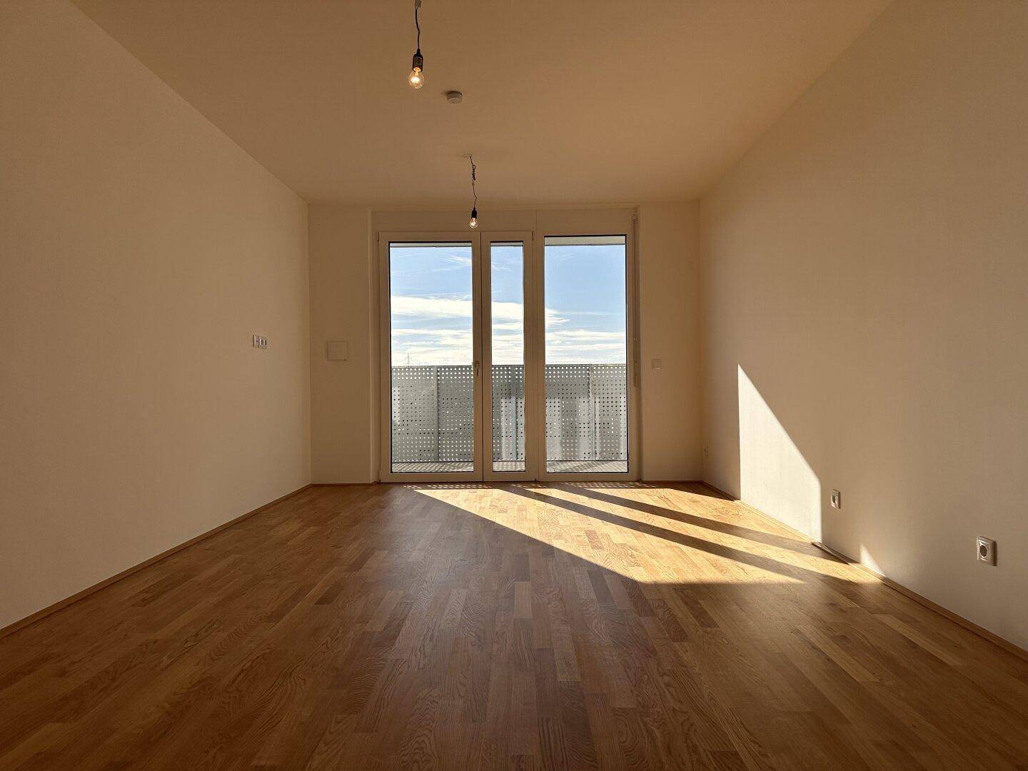 Großes, helles Zimmer mit Parkettboden und breiter Balkontür, durch die viel Tageslicht einfällt.