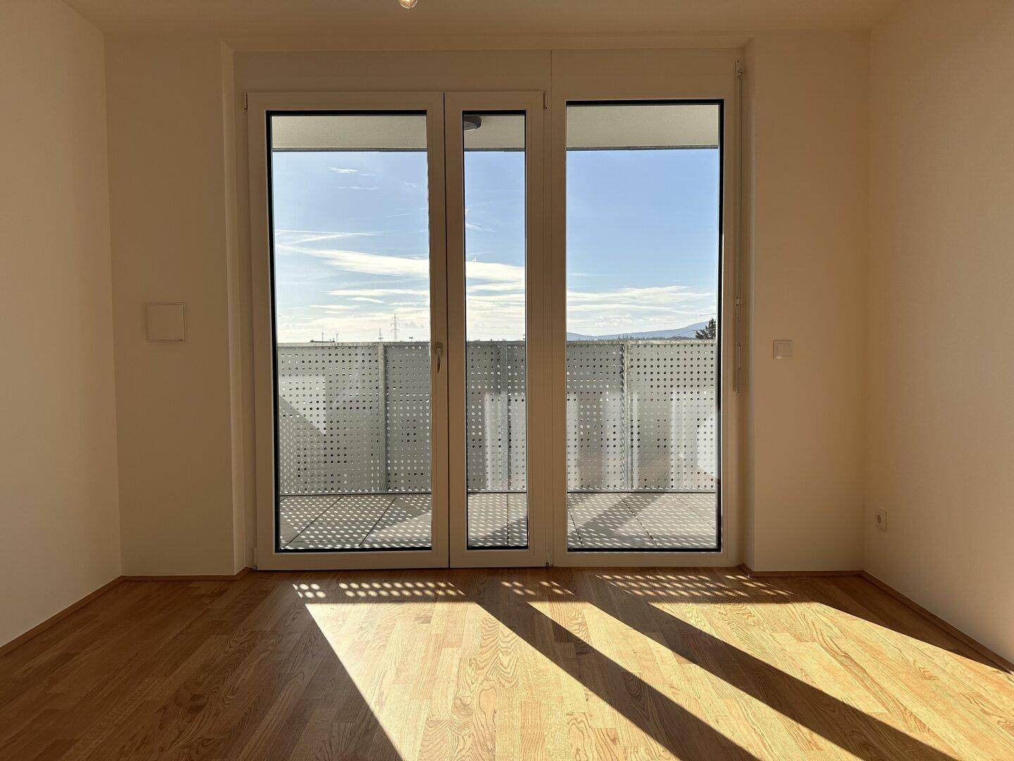 Helles Zimmer mit Parkettboden und Zugang zu einem Balkon, durchflutet von Sonnenlicht.