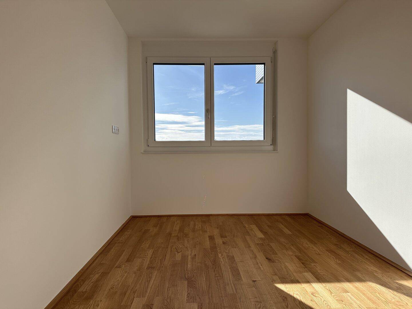 Helles Schlafzimmer mit Parkettboden und großem Fenster, das viel Tageslicht hereinlässt.