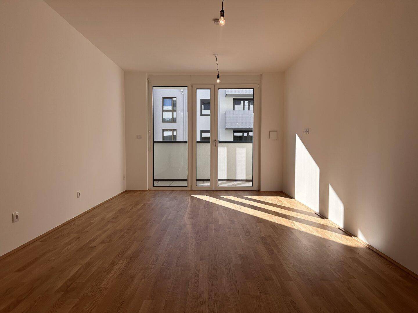 Helles, leeres Wohnzimmer mit Parkettboden und Zugang zu einem Balkon, durchflutet von Sonnenlicht.