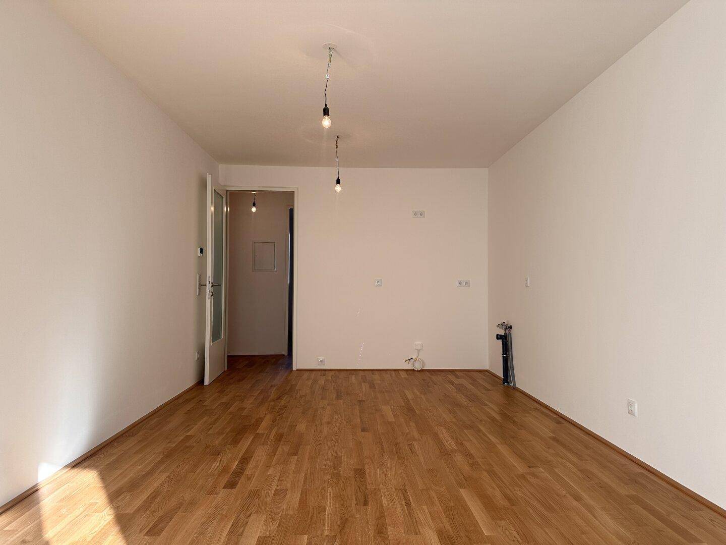 Geräumiges, leeres Zimmer mit Holzboden und Blick auf den Flur in einem Neubau.