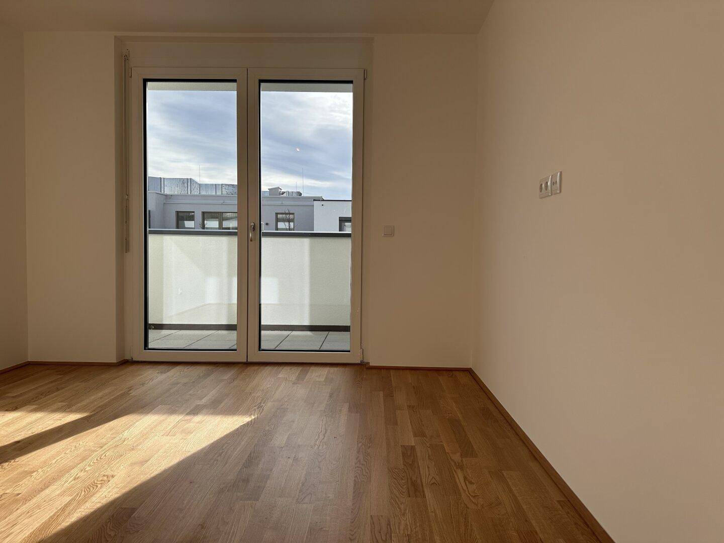 Lichtdurchfluteter Raum mit Parkettboden und breiter Balkontür, bietet viel Tageslicht.