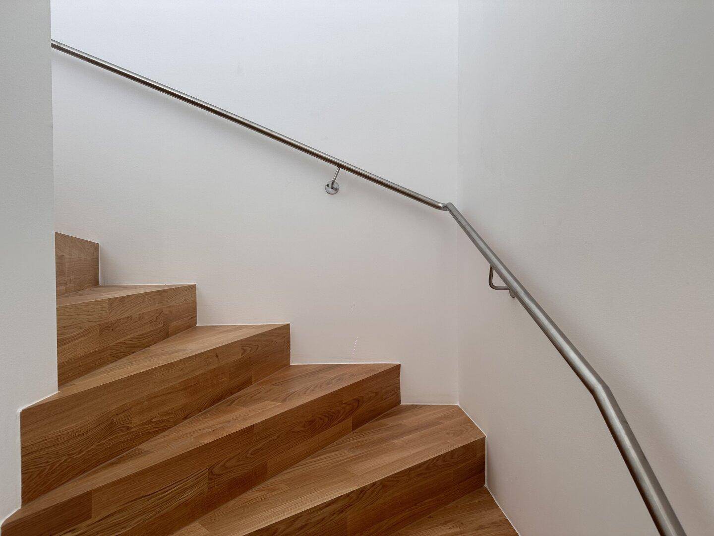Detailansicht der Holztreppe mit modernem Edelstahlgeländer in einem hellen Treppenhaus.