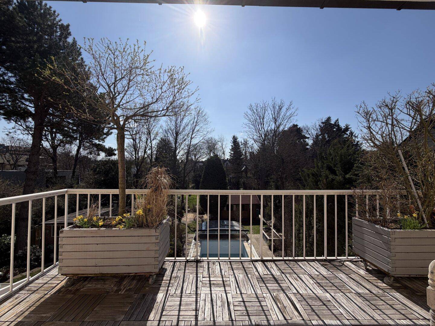 Sonniger Balkon mit Holzbelag und weitem Blick auf den Garten und den Poolbereich.