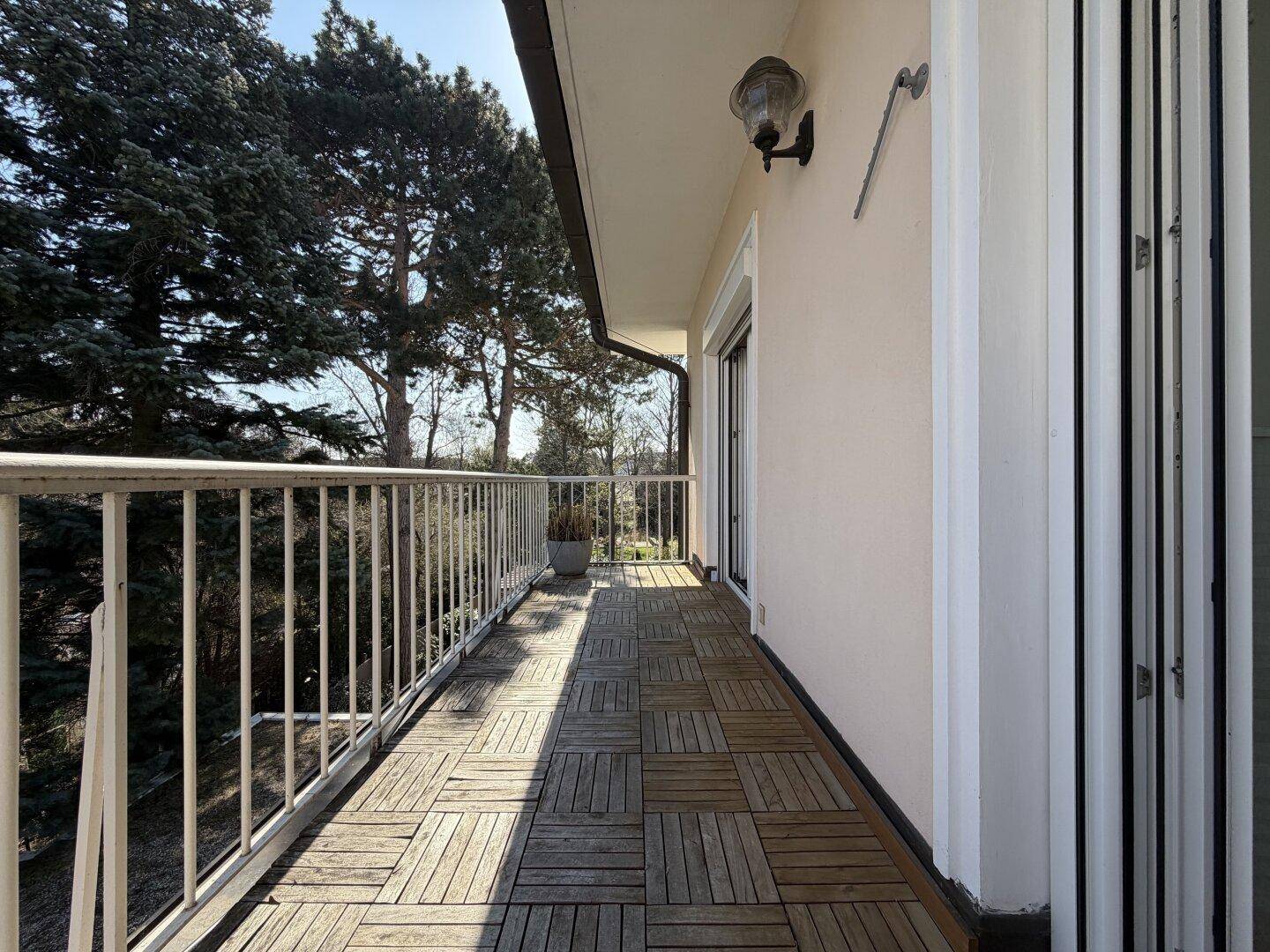 Langer Balkon mit Holzboden und weißem Geländer, umgeben von Bäumen für Privatsphäre.