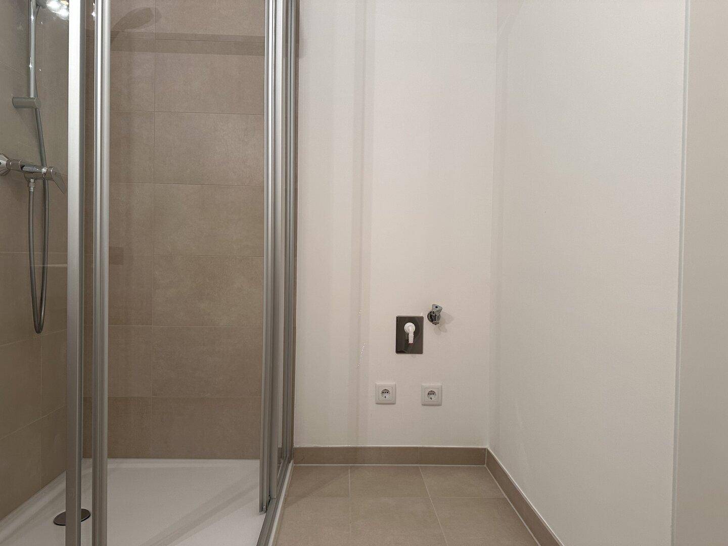 Duschbereich im Badezimmer mit beigefarbenen Fliesen und Anschluss für eine Waschmaschine.