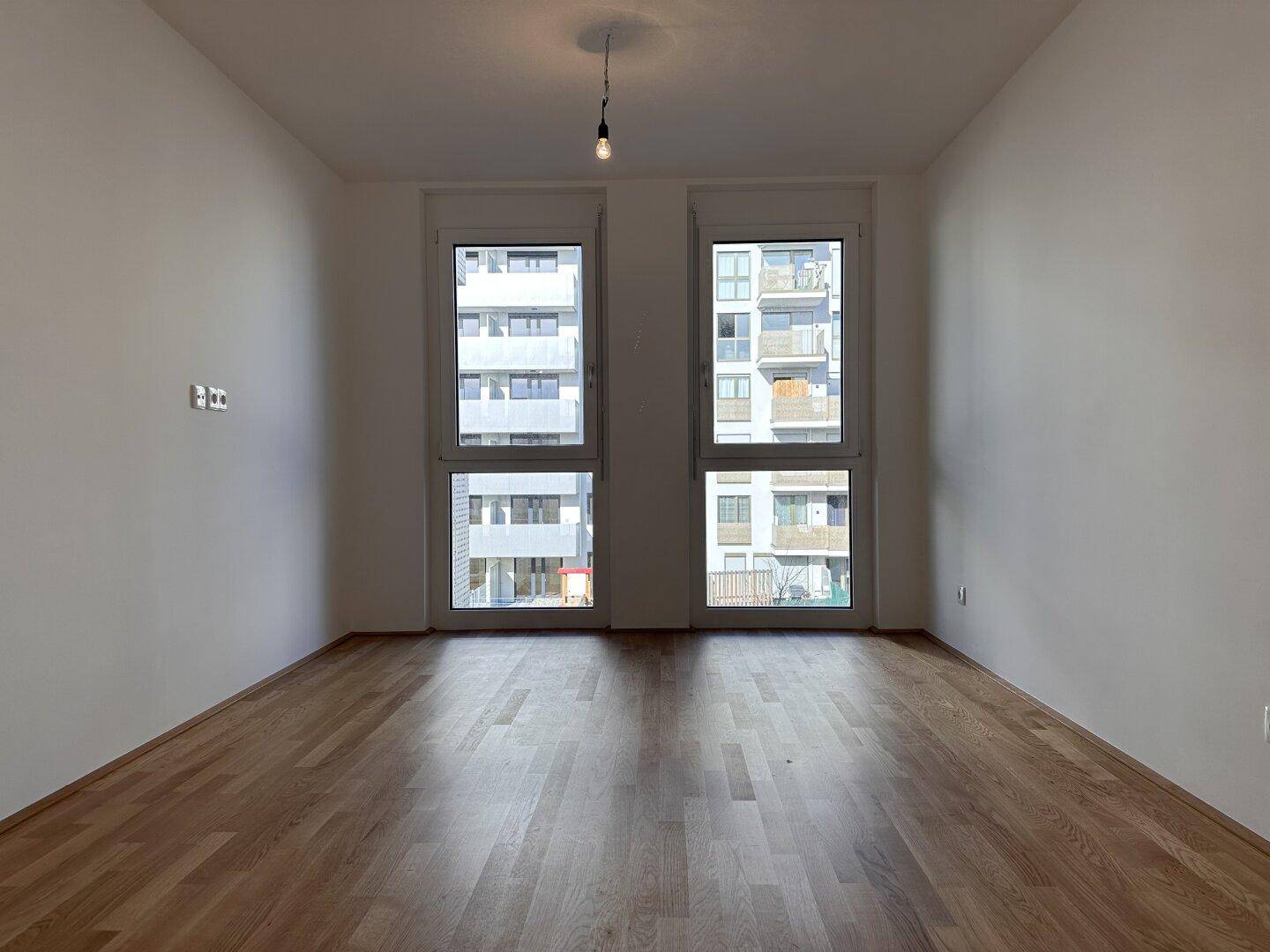 Helles Zimmer mit Parkettboden und großen Fenstern, die einen Blick auf die umliegenden Gebäude bieten.