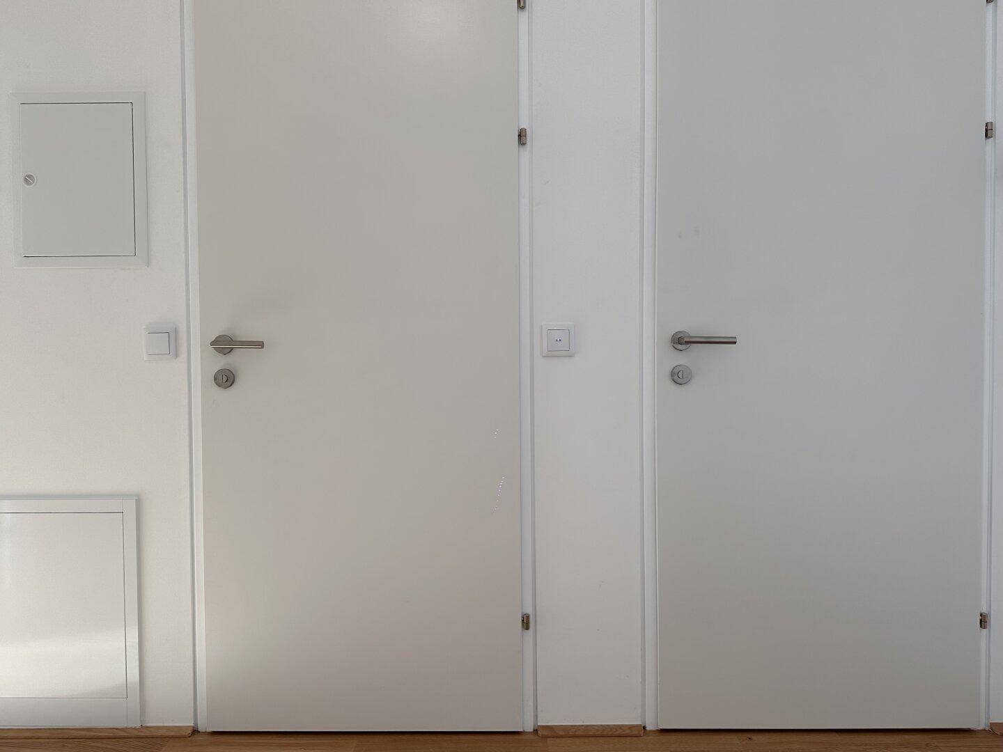 Detailansicht von zwei weißen Innentüren und einer Wand mit Wartungsklappen in einem hellen Flur.