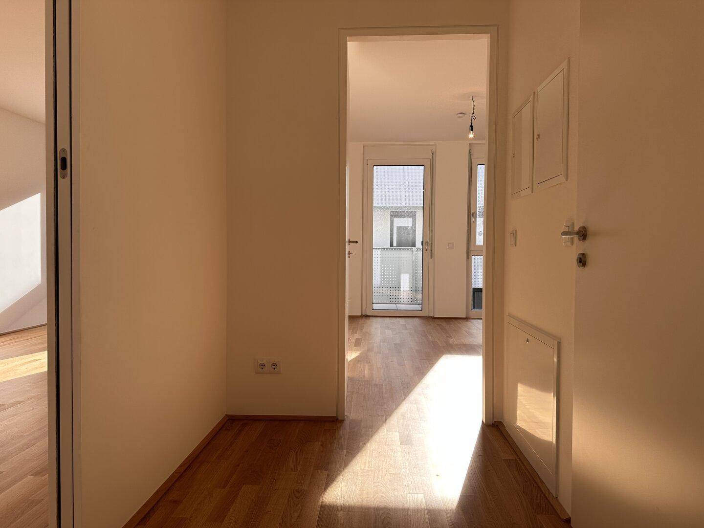 Langer Flur mit Holzboden, der in einen hellen Raum mit Fenstern und Zugang zum Außenbereich führt.