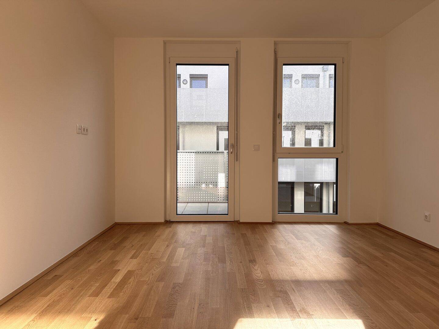 Geräumiges Zimmer mit Parkettboden und großen Fenstern, die viel Tageslicht hereinlassen und einen Blick nach draußen bieten.