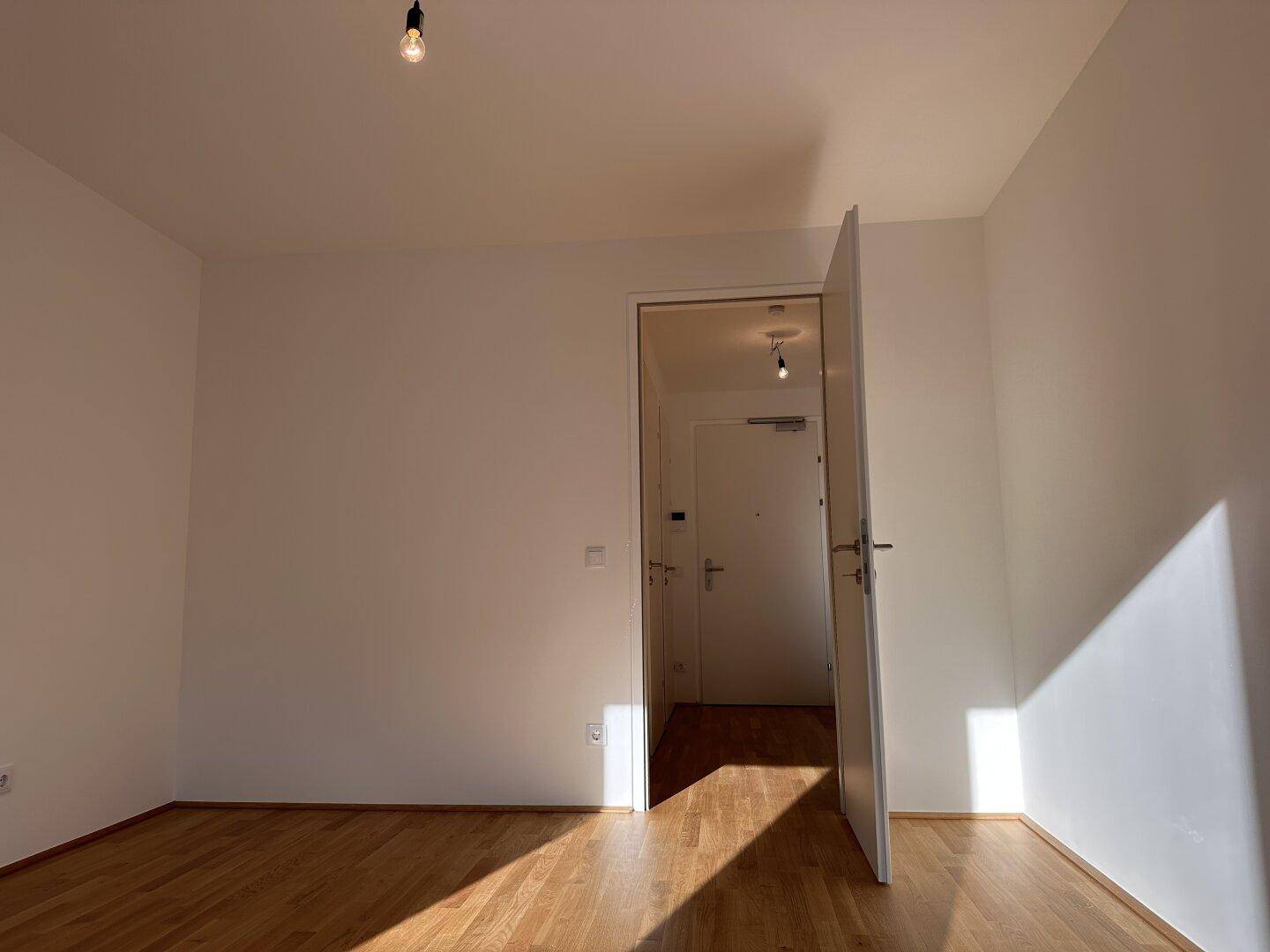 Helles Zimmer mit Parkettboden und offener Tür, die den Blick in den Flur freigibt, durchflutet von Sonnenlicht.