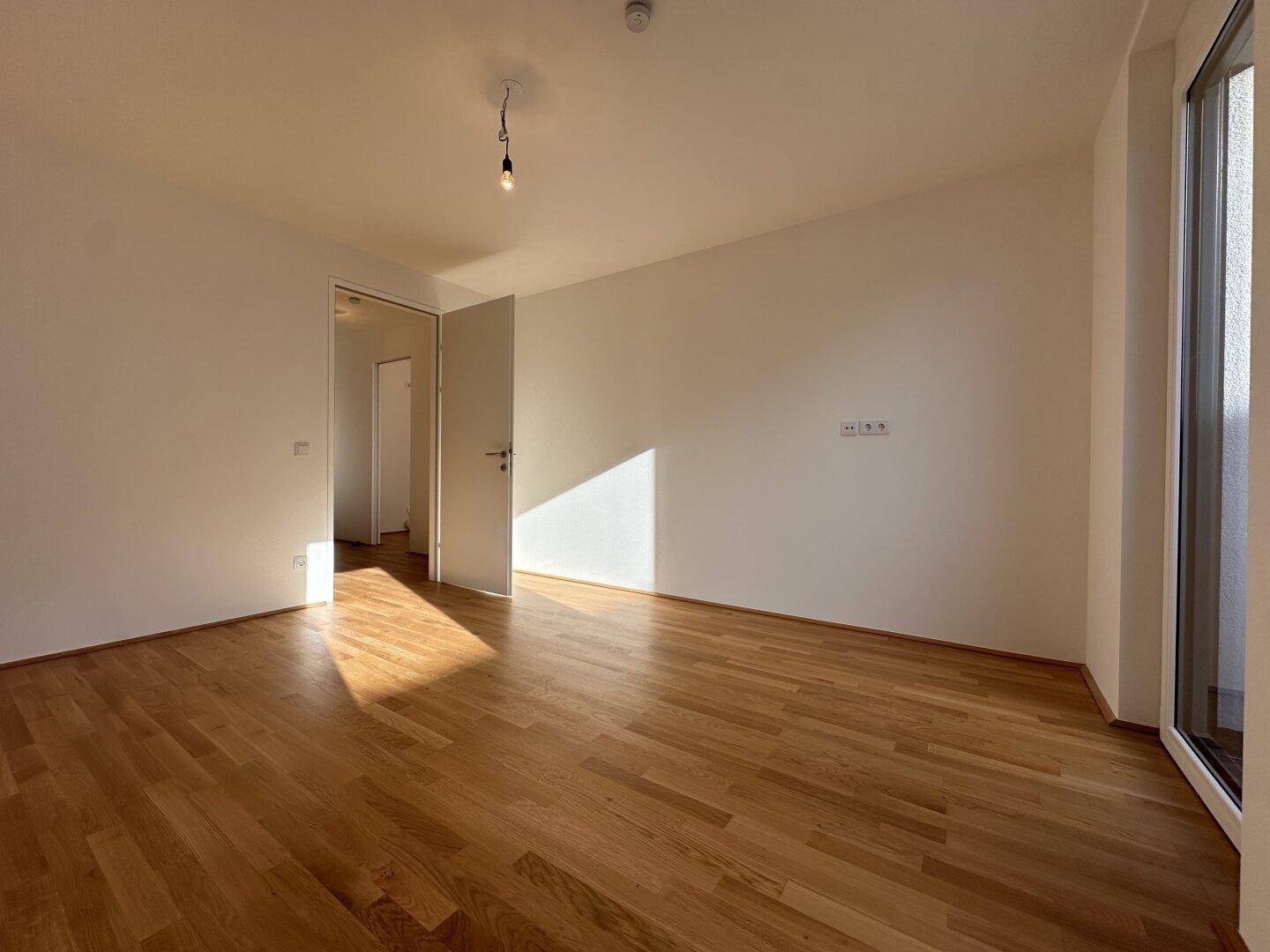 Großes, helles Zimmer mit Parkettboden und Zugang zu einem weiteren Raum, mit viel natürlichem Lichteinfall.