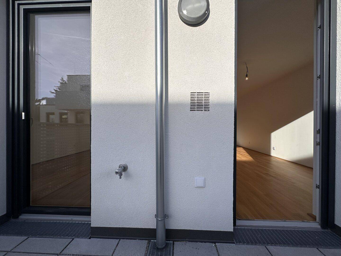 Außenwand mit Regenrohr, Wasserhahn und Lüftungsgitter neben einer großen Balkontür, die ins Innere führt.