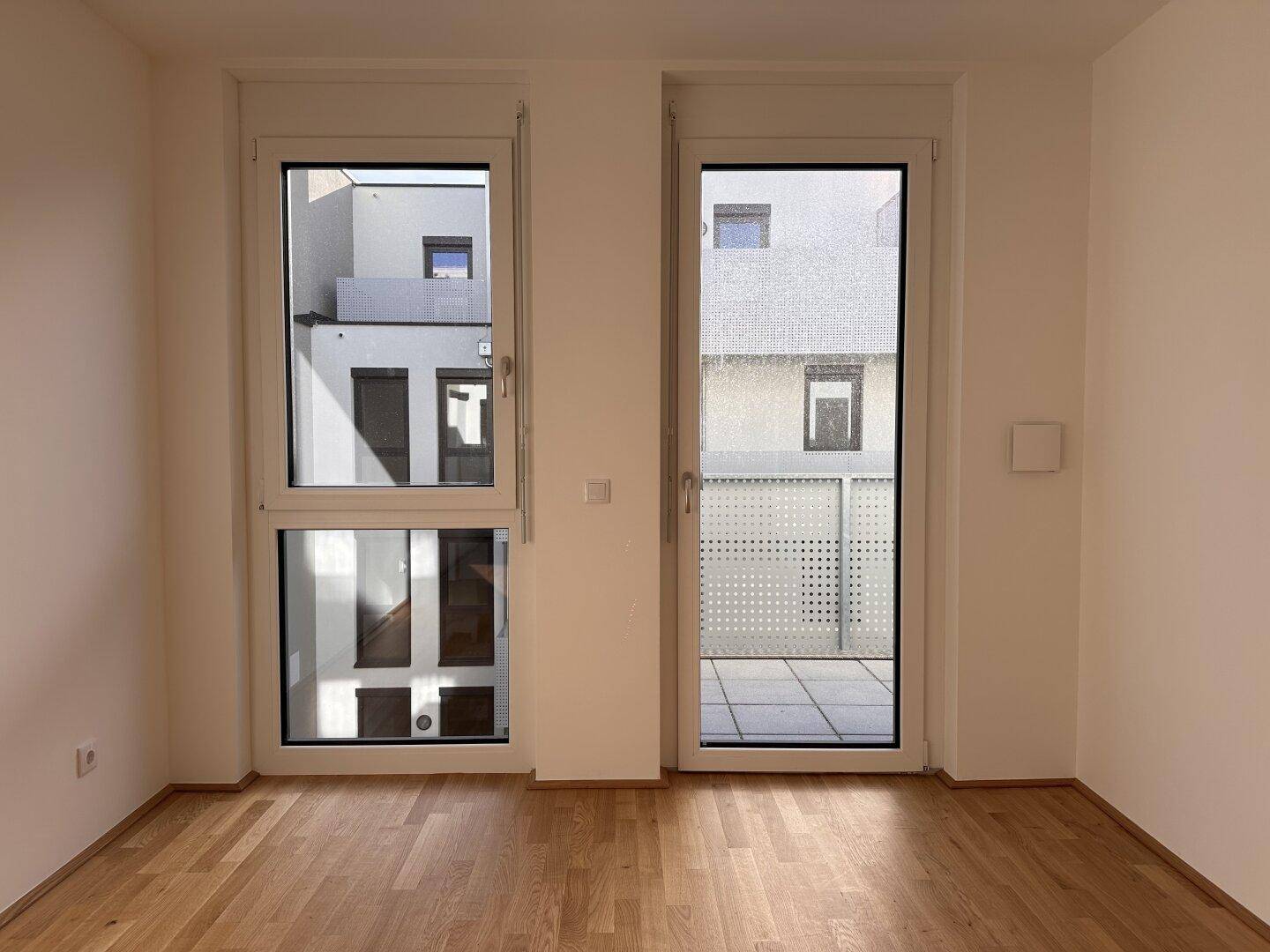 Helles Zimmer mit Parkettboden und großen Fenstern, die den Blick auf die moderne Gebäudefassade freigeben.