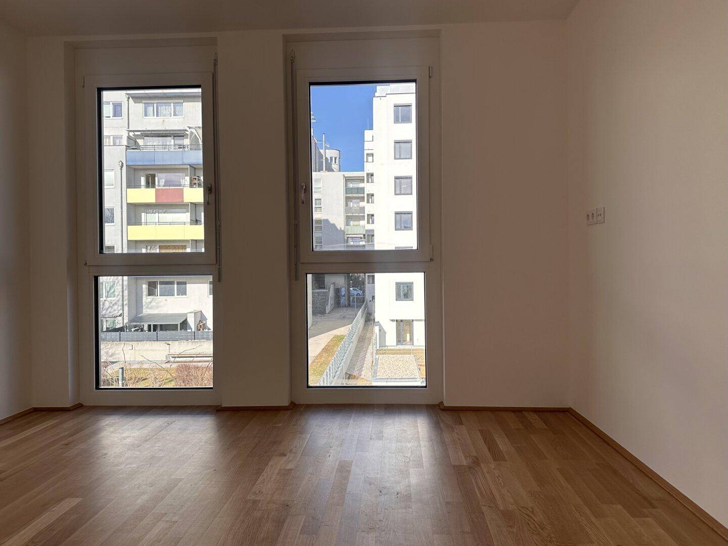 Helles Zimmer mit Parkettboden und großen Fenstern mit Blick auf die Umgebung.