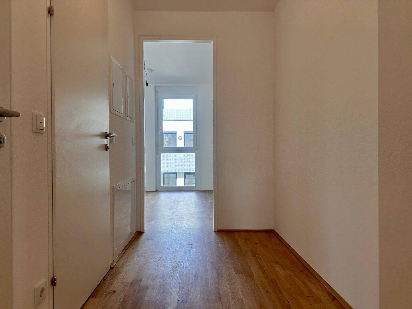 Schmaler Flur mit Holzboden und weiß gestrichenen Wänden, der zu einem hellen Raum führt.