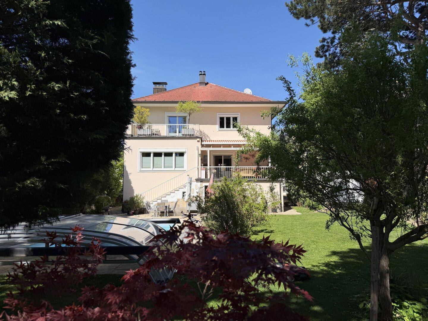 Hausfassade mit rotem Dach, umgeben von einem weitläufigen Garten und üppiger Vegetation.