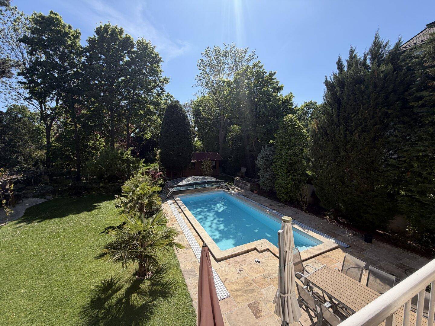 Panoramablick von oben auf den weitläufigen Garten mit Pool und umgebenden hohen Bäumen.