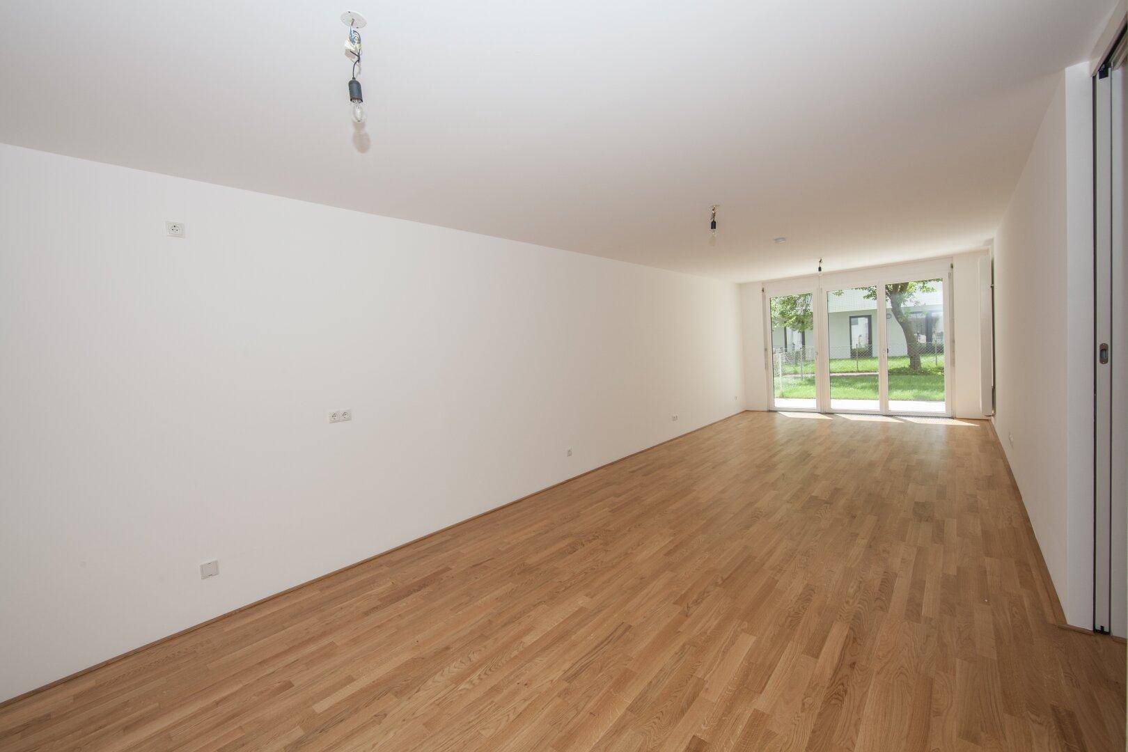Geräumiges, leeres Wohnzimmer mit hellem Parkettboden und großen Fenstern zum Garten.