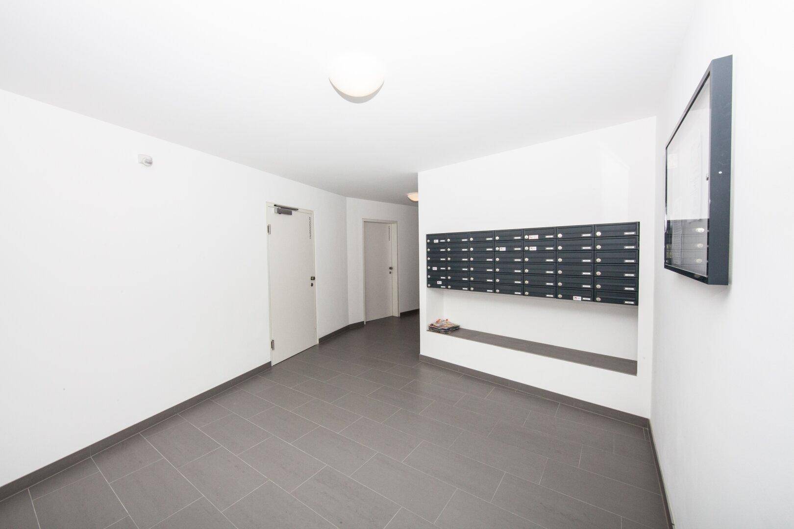 Heller Eingangsbereich mit grauen Fliesen und Briefkästen an der Wand in einem modernen Gebäude.