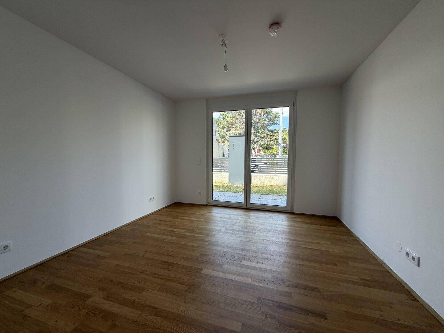 Geräumiges Zimmer mit Holzboden und großer Fenstertür, die viel Tageslicht hereinlässt.