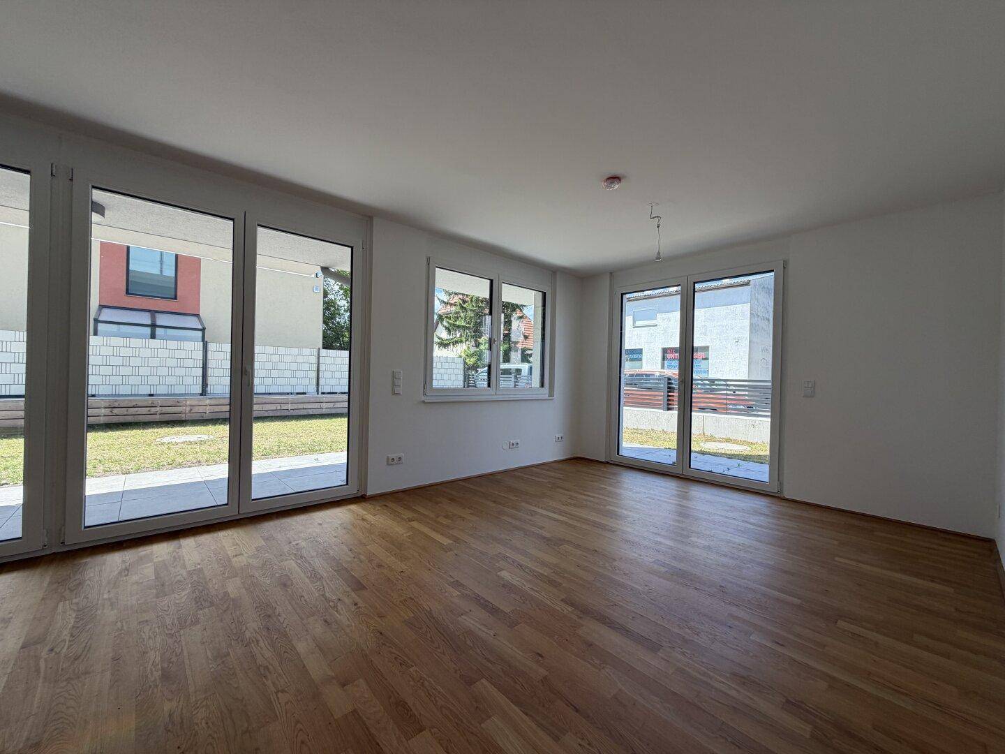 Heller, leerer Wohnraum mit großen Fenstern, Holzboden und Zugang zum Garten.
