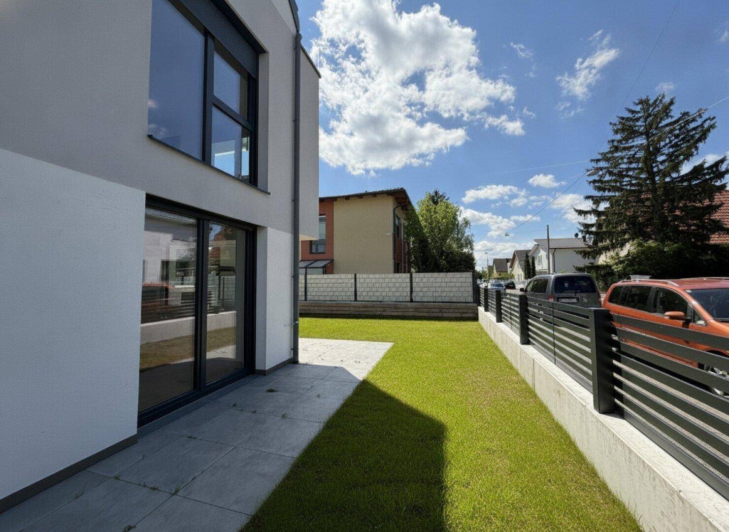 Gebäudeseite mit Terrasse, Rasenfläche und Blick auf die umliegende Wohngegend.